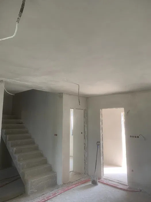 Surowe wnętrze w trakcie prac wykończeniowych: widoczne świeżo położone tynki na ścianach i suficie, betonowe schody, nieotynkowane ościeżnice drzwiowe oraz wystające przewody elektryczne.