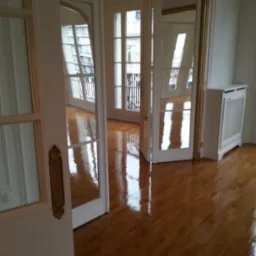 Błyszcząca, parkietowa podłoga w odnowionym pokoju z białymi ścianami, widoczne okna z balkonami i drzwi z ozdobnymi, złotymi klamkami.