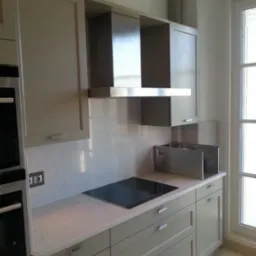 Nowoczesna kuchnia w odcieniach szarości z marmurowym blatem, płytą indukcyjną i metalowym okapem, oświetlona naturalnym światłem z okna.