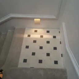 Schody wyłożone płytkami w biało-czarną szachownicę, oświetlone kwadratową lampą sufitową, widok z góry.