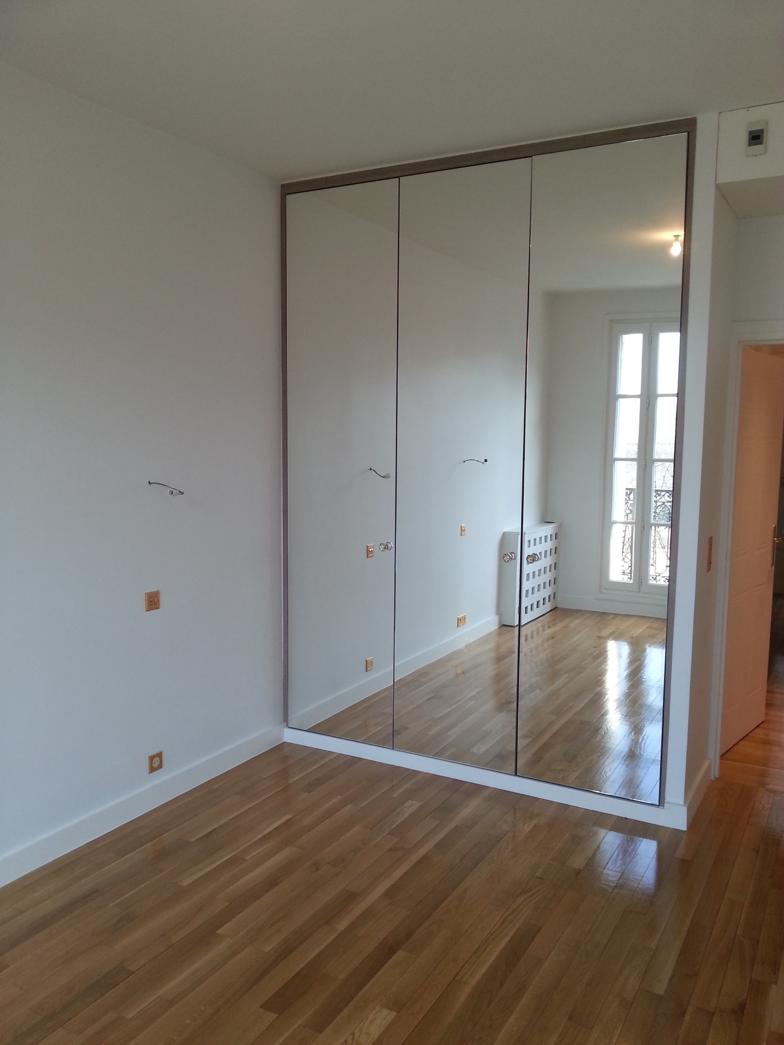 Wnękowa szafa z lustrzanymi drzwiami przesuwnymi, odbijająca parkiet i okno w jasnym pokoju.