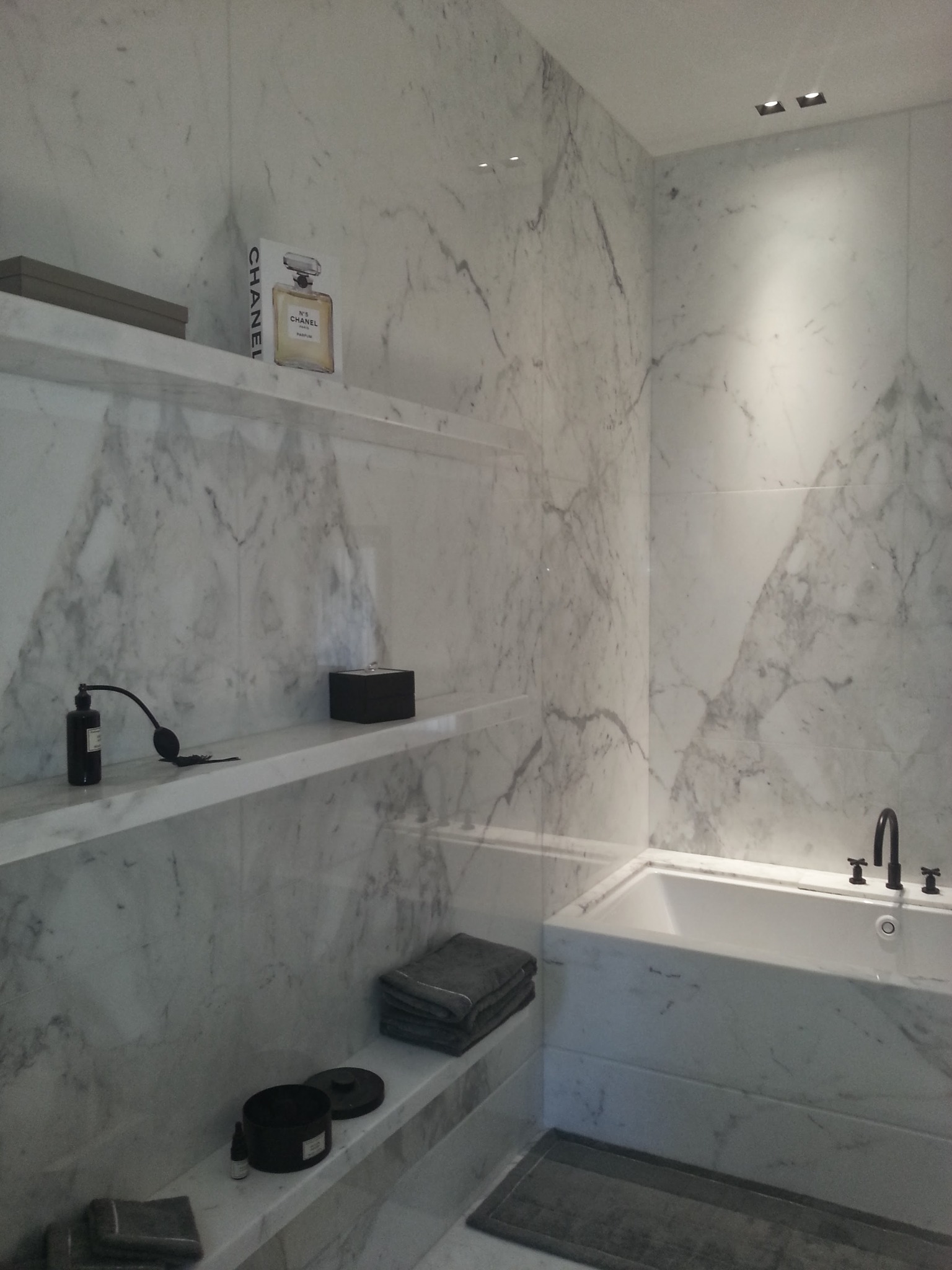 Luksusowa łazienka wykończona marmurem Carrara, z półkami, na których stoją ozdobne flakony perfum i kosmetyki. Widoczna wanna z czarną armaturą i szare ręczniki.
