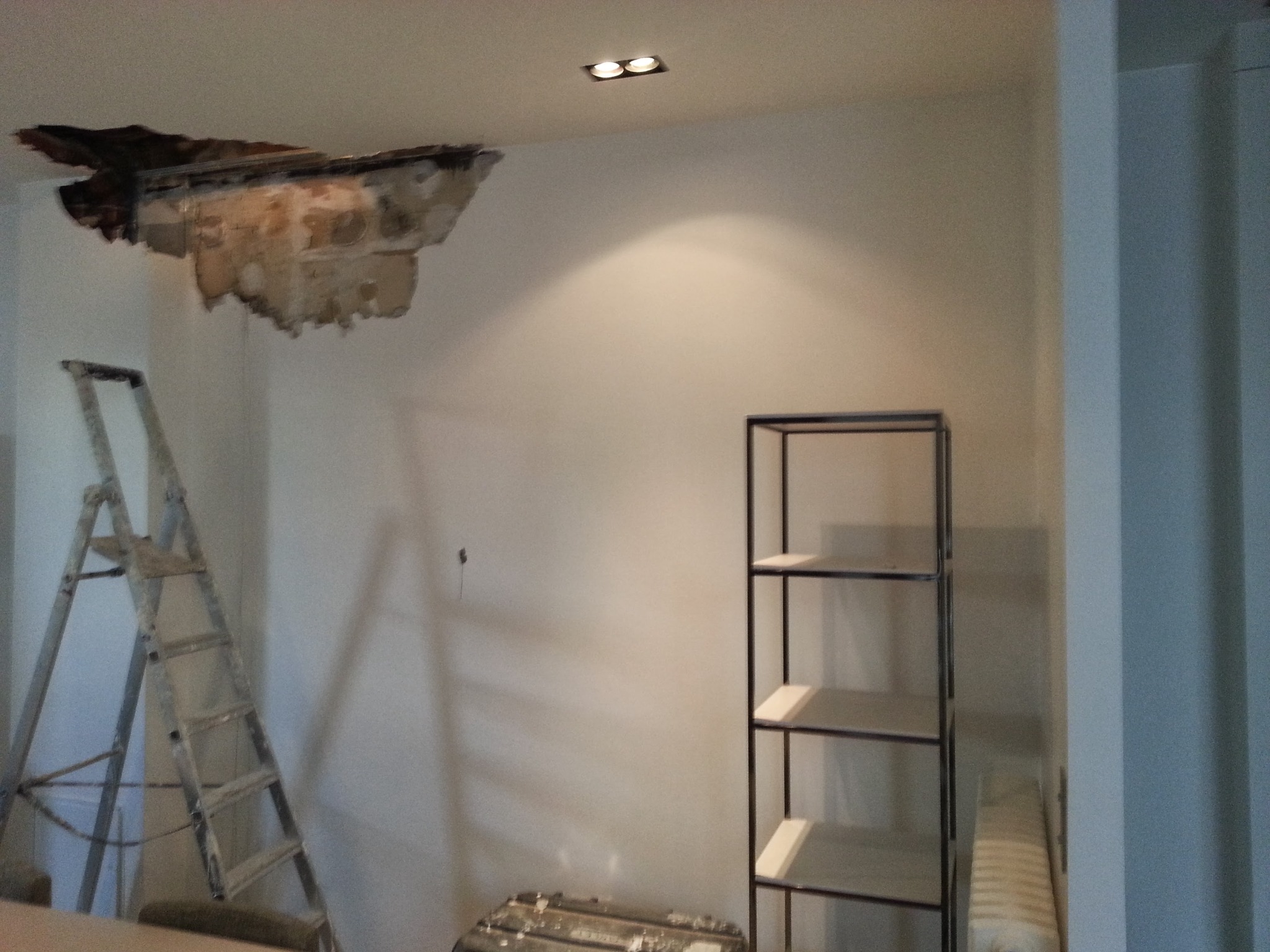 Uszkodzony sufit z widocznymi warstwami, drabina oparta o ścianę, półka z metalową ramą, przygotowania do naprawy sufitu.
