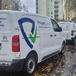 Dwa białe busy firmy ProperGuard z logo i danymi kontaktowymi zaparkowane na mokrej ulicy w jesienny dzień. Oferują doradztwo, projekt, instalację i serwis systemów bezpieczeństwa.