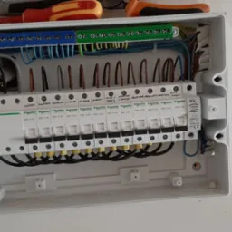 Otwarta skrzynka rozdzielcza z zainstalowanymi bezpiecznikami Schneider Electric, widoczne kable w różnych kolorach oraz listwy zaciskowe, narzędzia u góry obudowy.