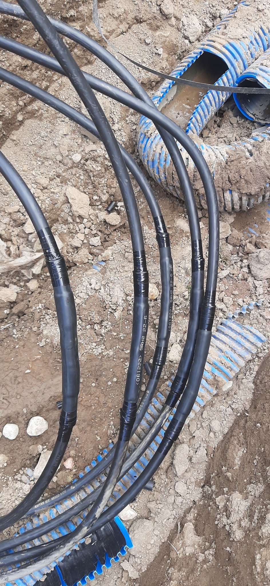Ułożone w ziemi kable elektryczne w czarnej izolacji, połączone taśmą izolacyjną, leżące obok niebieskiej rury karbowanej.