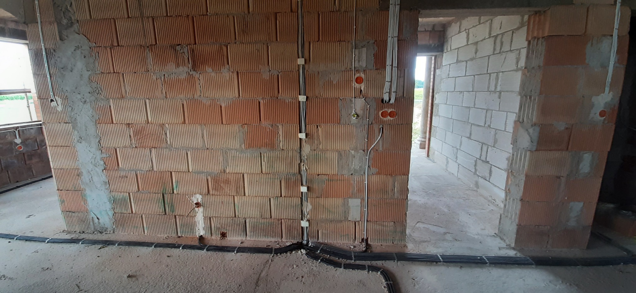 Wewnętrzne ściany z czerwonej cegły z widocznymi przewodami elektrycznymi w peszlach prowadzonymi wzdłuż podłogi i pionowo po ścianach, przygotowane pod tynk.