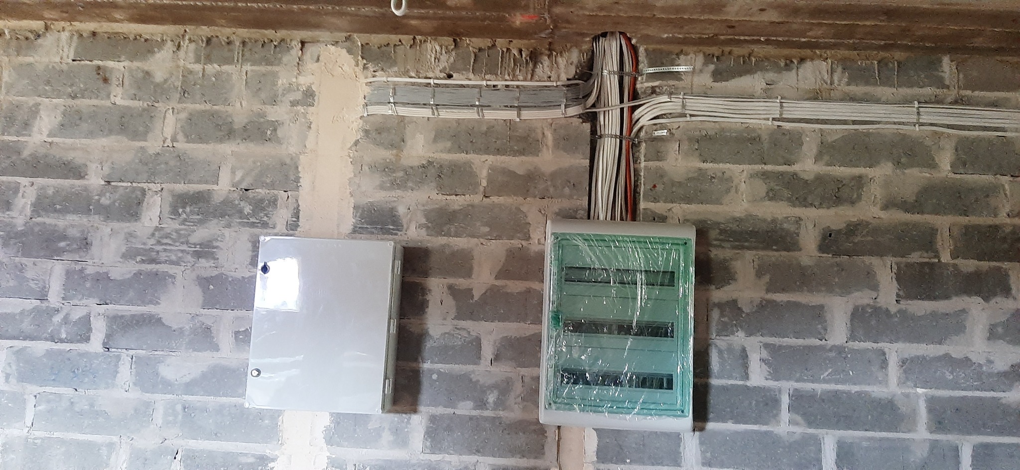 Dwa panele elektryczne zamontowane na surowej ścianie z betonowych pustaków. Jeden panel jest biały, drugi zabezpieczony folią. Nad nimi widoczne kable elektryczne poprowadzone w korytkach.