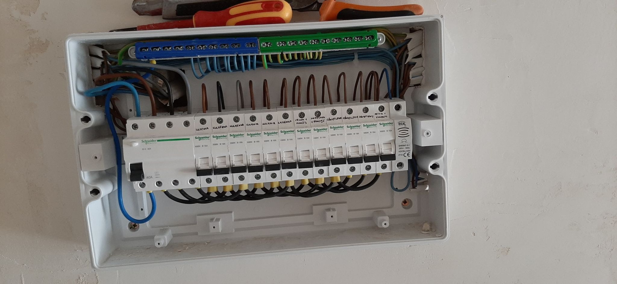 Otwarta skrzynka rozdzielcza z zainstalowanymi bezpiecznikami Schneider Electric, widoczne kable w różnych kolorach oraz listwy zaciskowe, narzędzia u góry obudowy.