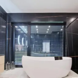 Nowoczesna łazienka z czarnymi marmurowymi płytkami, wolnostojącą białą wanną i oknem z żaluzjami, oświetlona punktowymi lampami sufitowymi.