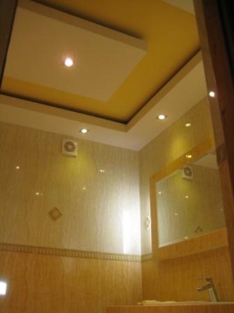 Wykończona łazienka z podwieszanym sufitem w kolorze żółtym i białym, oświetleniem punktowym oraz marmurowymi płytkami na ścianach i podłodze, widoczne lustro i umywalka z baterią.