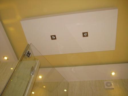 Podwieszany sufit w łazience z dwoma kwadratowymi lampami wpuszczanymi, widoczna kabina prysznicowa z przezroczystego szkła.