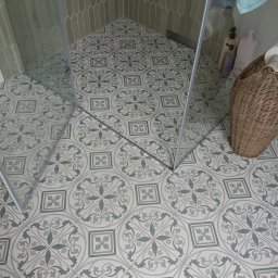 OLEKSANDR BEZOTOSNYI - Wykończona łazienka z kabiną prysznicową. Na podłodze i ścianach wzorzyste, szaro-zielone płytki. W rogu kosz wiklinowy z akcesoriami. Widok z góry.