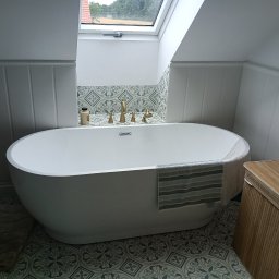 OLEKSANDR BEZOTOSNYI - Biała wanna wolnostojąca w łazience na poddaszu z oknem dachowym. Ściany wyłożone panelami i dekoracyjnymi płytkami. Złota armatura, ręcznik w paski i bambusowy kosz na pranie.