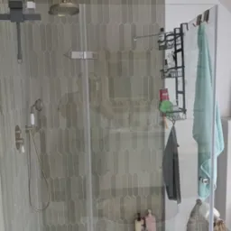 Szczegółowe ujęcie kabiny prysznicowej z armaturą w odcieniach złota i szaro-zielonymi, podłużnymi płytkami. Na podłodze wzorzyste płytki.