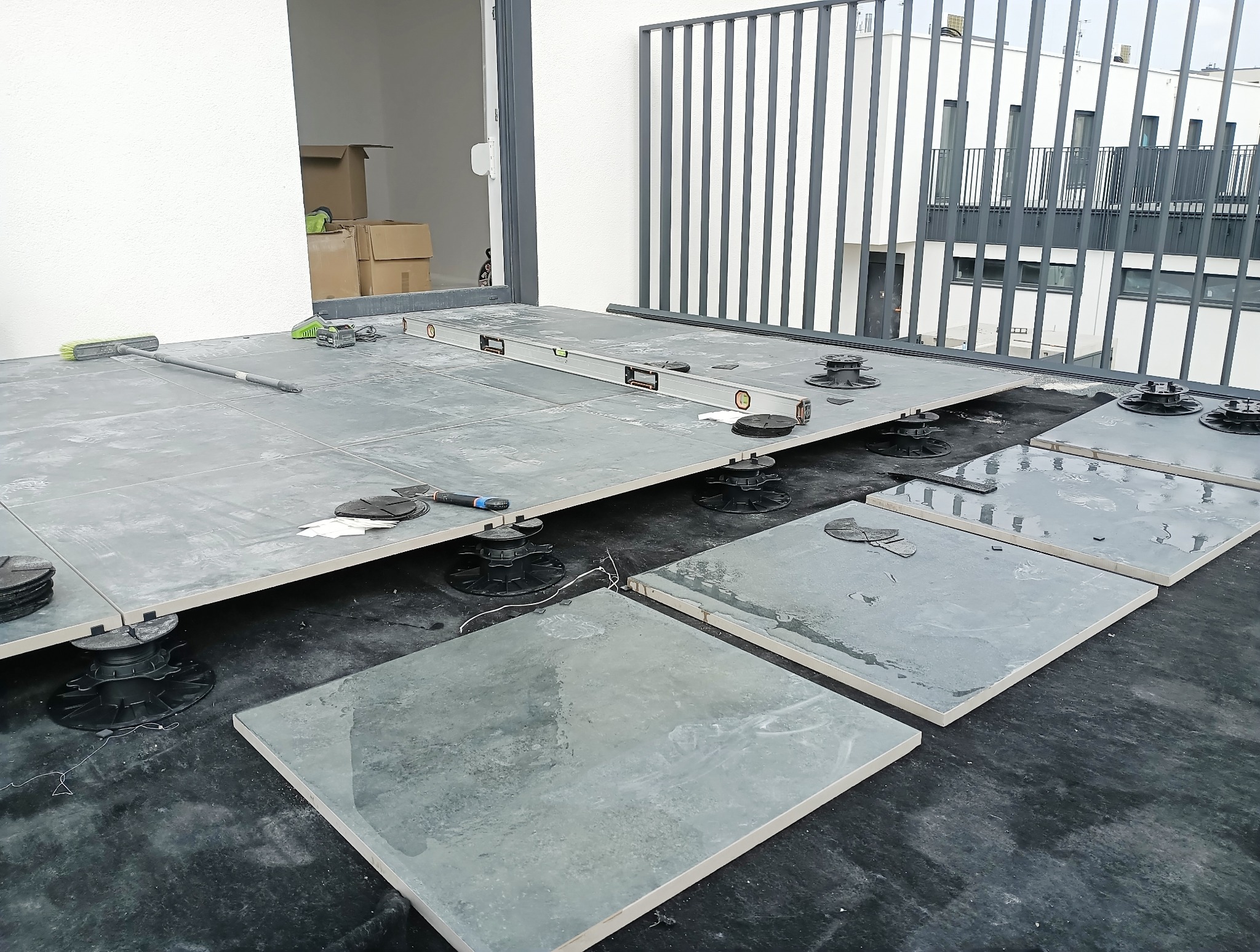 Montaż płytek na regulowanych wspornikach na tarasie. Widoczny poziomica, szczotka i narzędzia. Częściowo ułożone płytki. Tło: budynek mieszkalny i barierka.