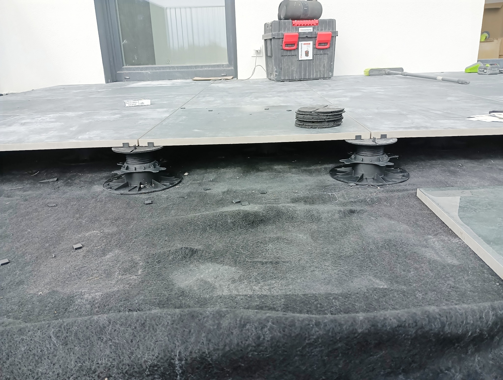 Montaż szarych płyt tarasowych na regulowanych wspornikach, widoczne narzędzia i akcesoria. Etap prac na zewnątrz budynku.