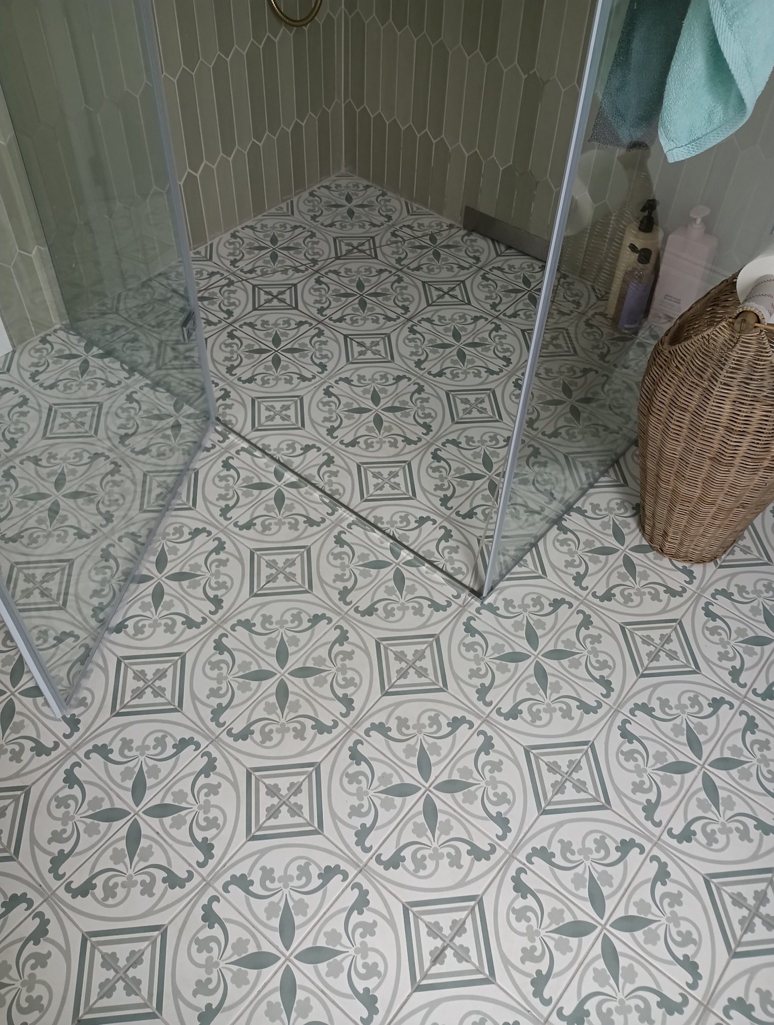 Wykończona łazienka z kabiną prysznicową. Na podłodze i ścianach wzorzyste, szaro-zielone płytki. W rogu kosz wiklinowy z akcesoriami. Widok z góry.