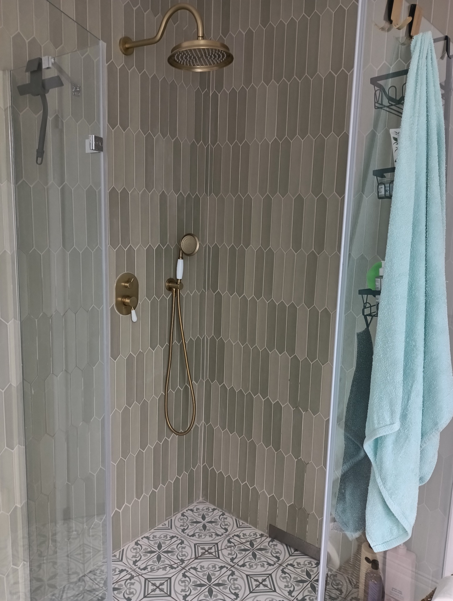 Kabina prysznicowa z szarą, pionową mozaiką w kształcie migdałów, złotą armaturą i wzorzystą podłogą. Na szklanej ściance wisi turkusowy ręcznik.