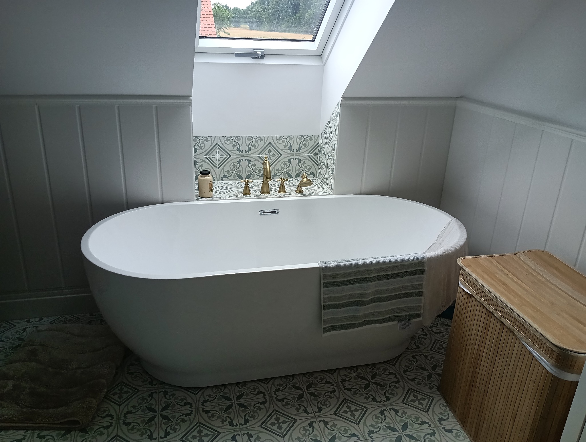 Biała wanna wolnostojąca w łazience na poddaszu z oknem dachowym. Ściany wyłożone panelami i dekoracyjnymi płytkami. Złota armatura, ręcznik w paski i bambusowy kosz na pranie.