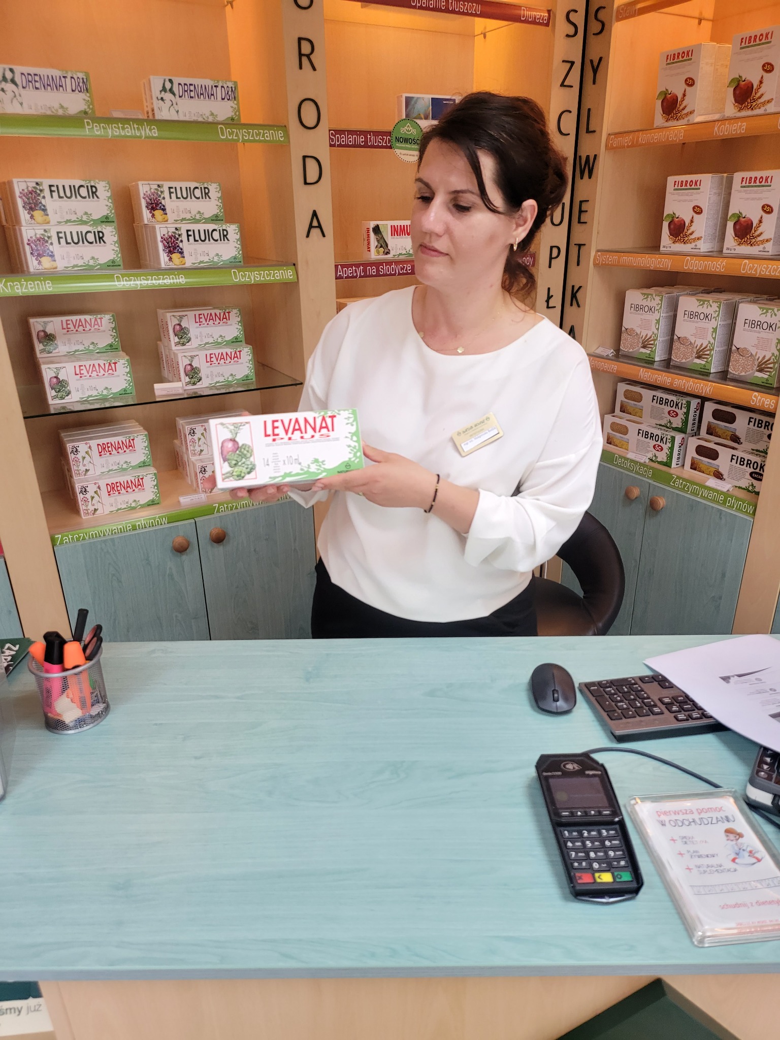 Sprzedawczyni w białej bluzce prezentuje opakowanie suplementu diety 'Levanat Plus' na tle półek z innymi produktami zdrowotnymi w sklepie.