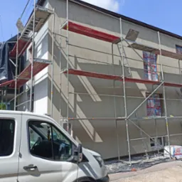 Budynek w trakcie ocieplania elewacji, widoczne rusztowania, zabezpieczone okna oraz częściowo otynkowana ściana, na pierwszym planie zaparkowany biały bus.