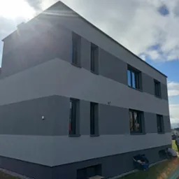 Dwukolorowa elewacja budynku z widocznymi pracownikami przy gruncie, błękitne niebo z chmurami w tle.