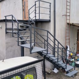 Artur Reszka Usługi ślusarskie i spawalnicze - Zewnętrzne schody metalowe z platformą i poręczami, prowadzące do drzwi na piętrze. Konstrukcja pomalowana na szaro, z kratowymi stopniami. W tle surowa elewacja budynku.