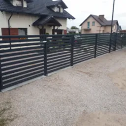 Nowoczesne, poziome ogrodzenie panelowe w kolorze antracytowym przed domem jednorodzinnym z szarą kostką brukową na podjeździe i otwartym bagażnikiem samochodu w tle.