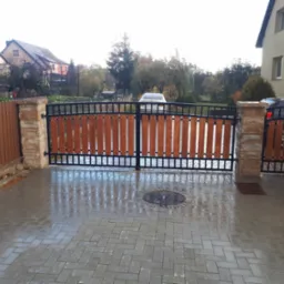 Dwuskrzydłowa brama wjazdowa z drewnianym wypełnieniem i czarną metalową ramą, osadzona na kamiennych słupkach, widoczna na mokrym brukowanym podjeździe.