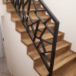 Drewniane schody z nowoczesną, geometryczną balustradą w kolorze grafitowym, zabezpieczoną taśmą na dole. Widoczne jasne ściany.