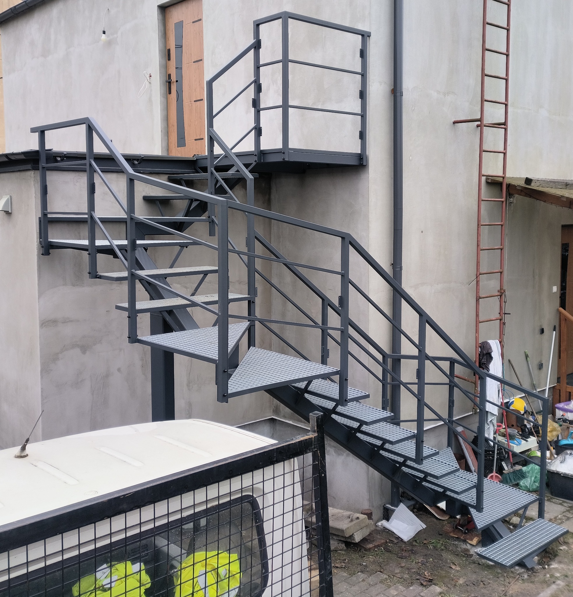 Zewnętrzne schody metalowe z platformą i poręczami, prowadzące do drzwi na piętrze. Konstrukcja pomalowana na szaro, z kratowymi stopniami. W tle surowa elewacja budynku.