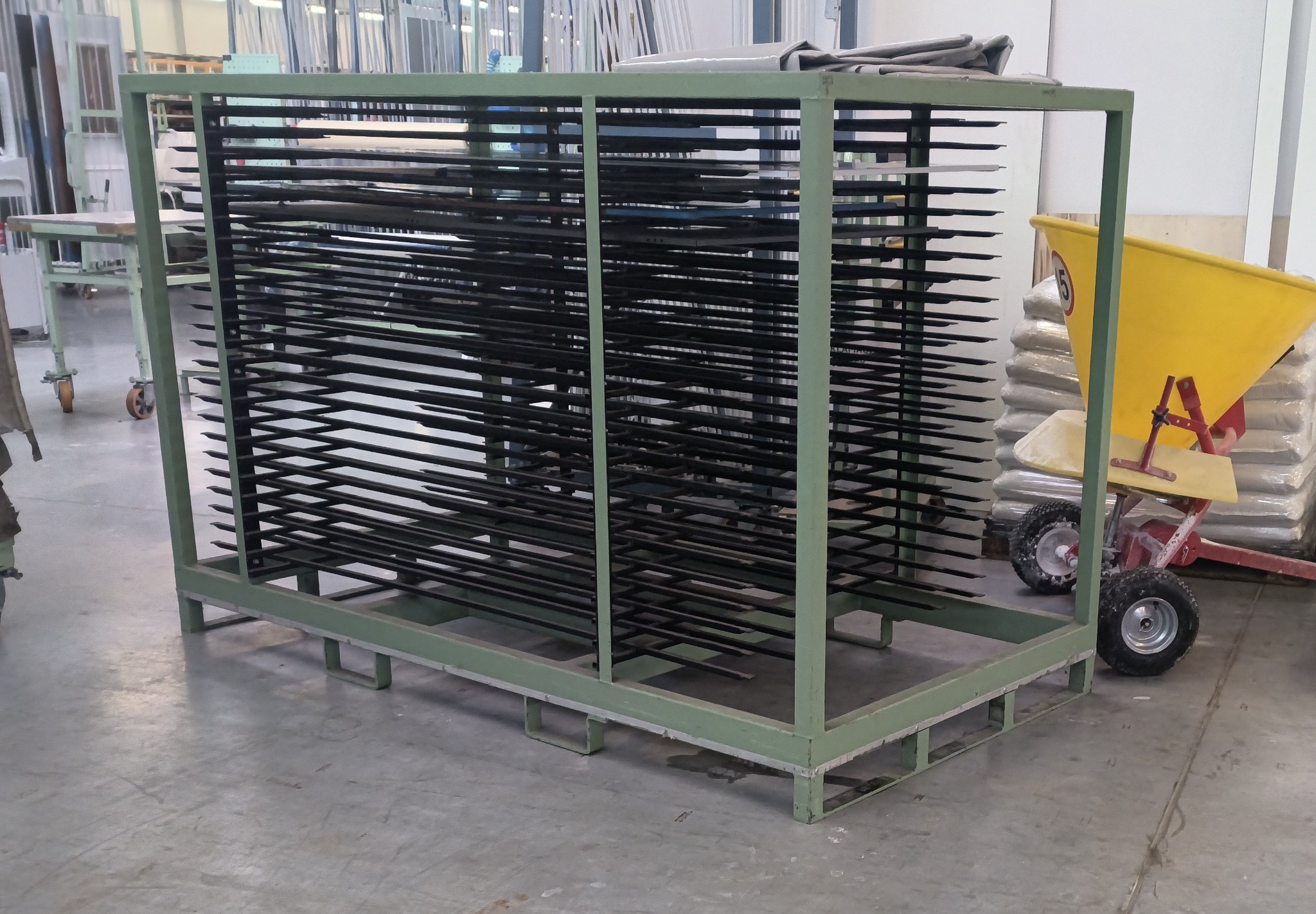 Metalowy regał z poziomymi prętami, prawdopodobnie do przechowywania elementów metalowych po obróbce, w tle widoczny wózek i materiały budowlane.