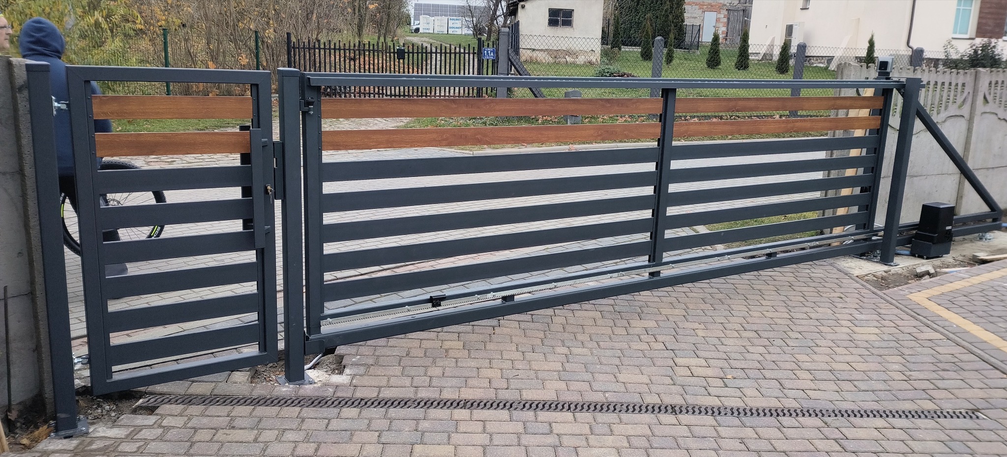Przesuwna brama wjazdowa z szarych, metalowych profili z drewnianymi akcentami, widoczny automat do bramy. Brama na posesji wyłożonej kostką brukową.
