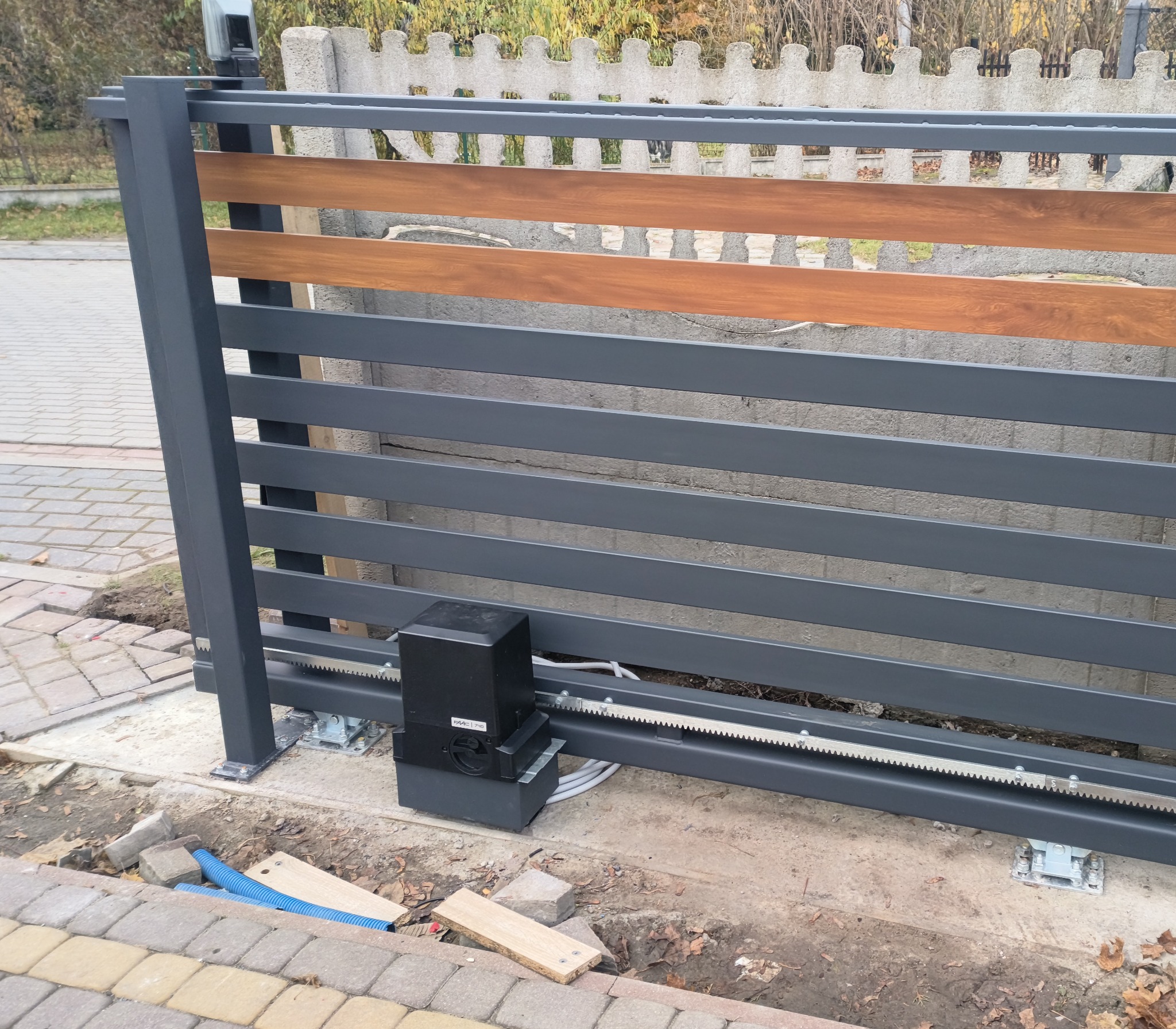 Nowoczesna brama wjazdowa przesuwna z szarymi, poziomymi panelami i drewnopodobnymi akcentami. Widoczny mechanizm automatycznego otwierania i fragment kostki brukowej.