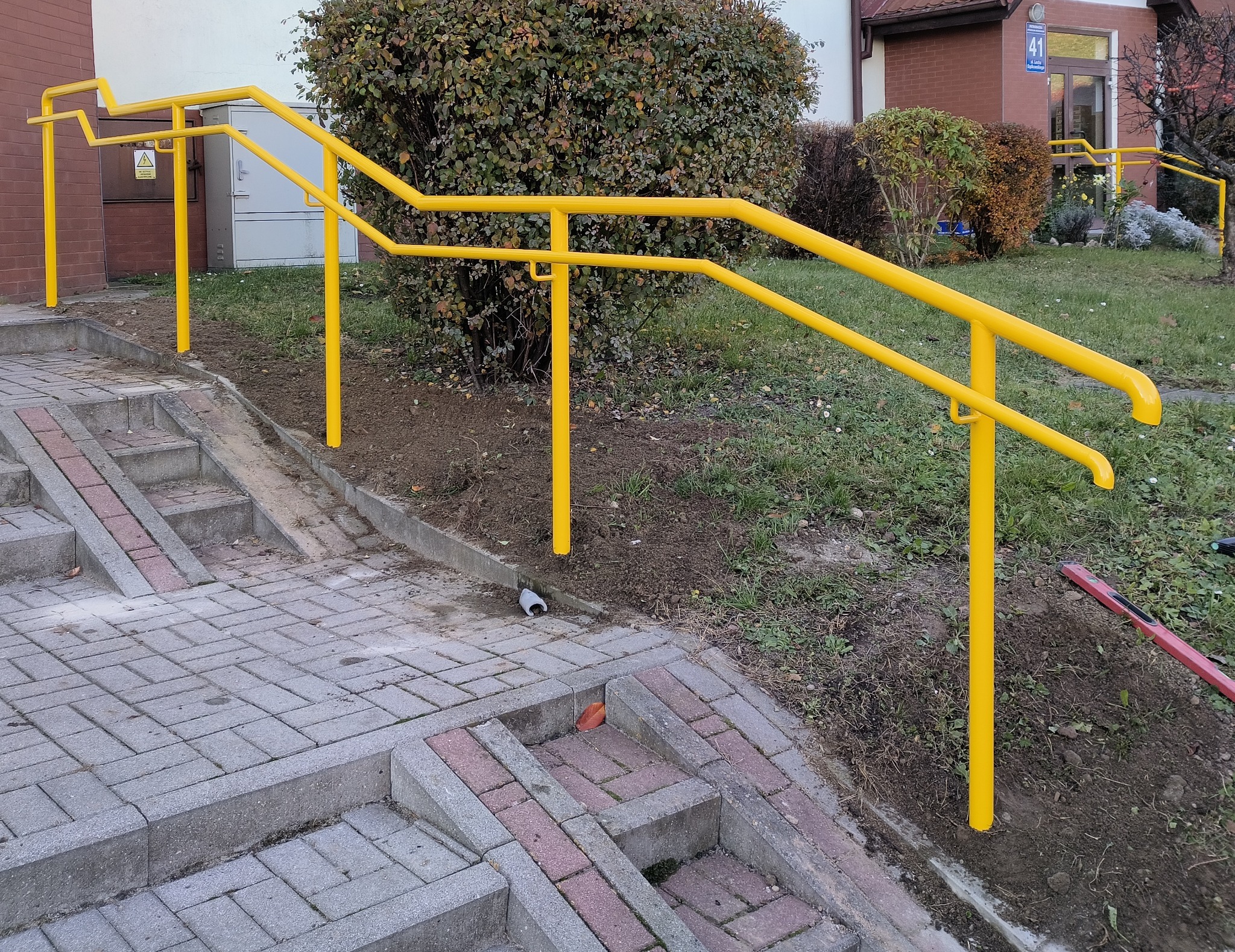 Żółte, metalowe poręcze przy schodach zewnętrznych z kostki brukowej, z rampą dla wózków. Tło stanowi budynek z numerem 41 i krzewy.