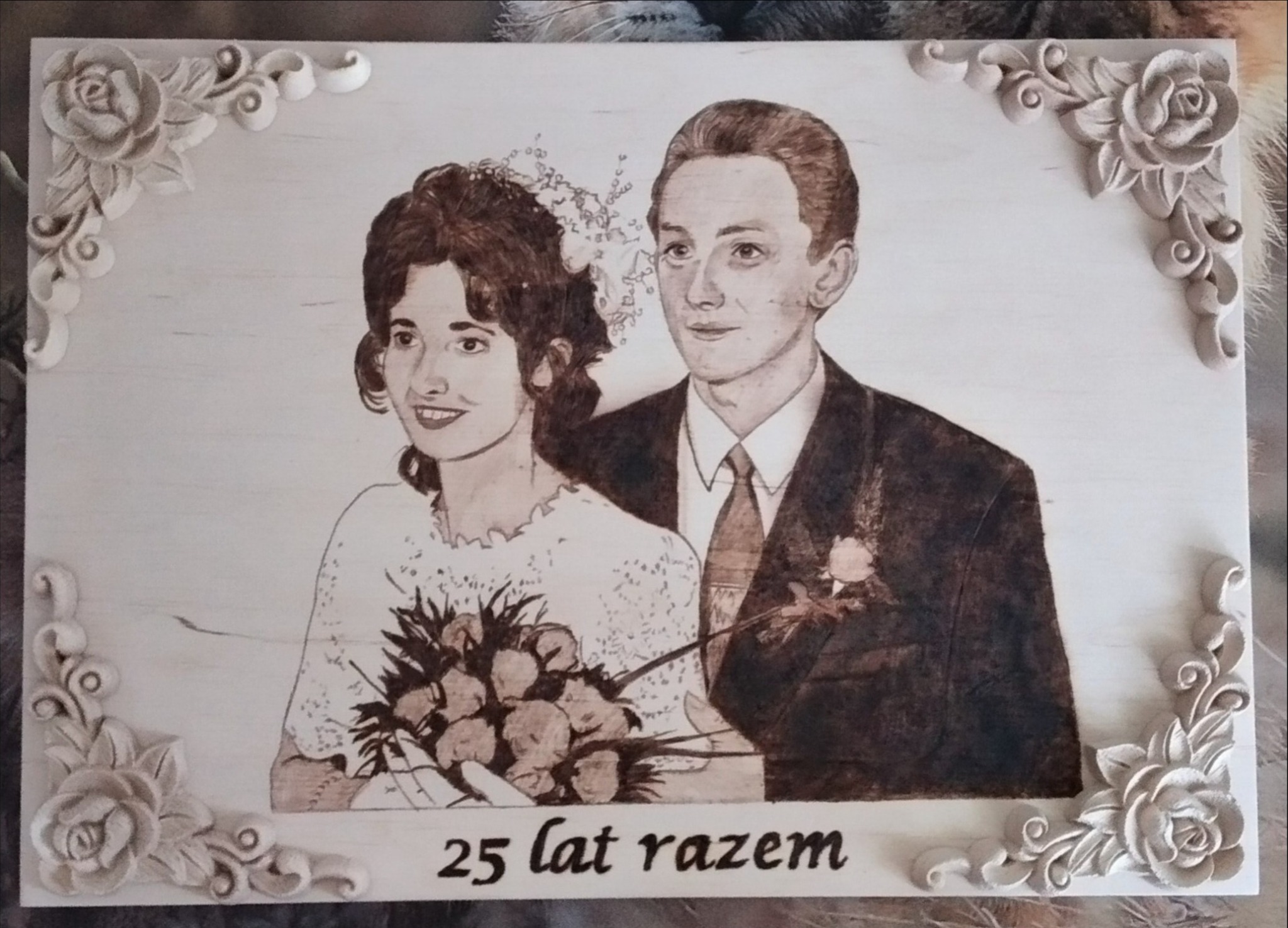Wypalony w drewnie portret ślubny pary z okazji 25 rocznicy, ozdobiony rzeźbionymi kwiatami w rogach.