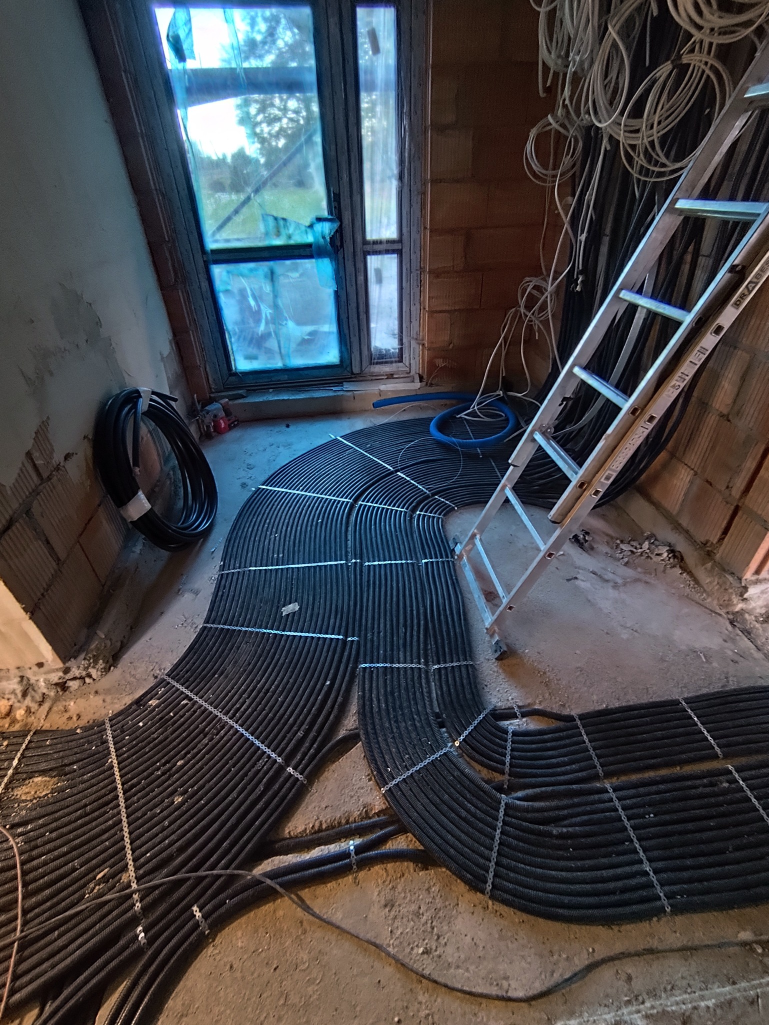 Układanie czarnych rur instalacji grzewczej podłogowej, mocowanych metalowymi klamrami, w surowym wnętrzu z oknem zabezpieczonym folią i drabiną opartą o ścianę.