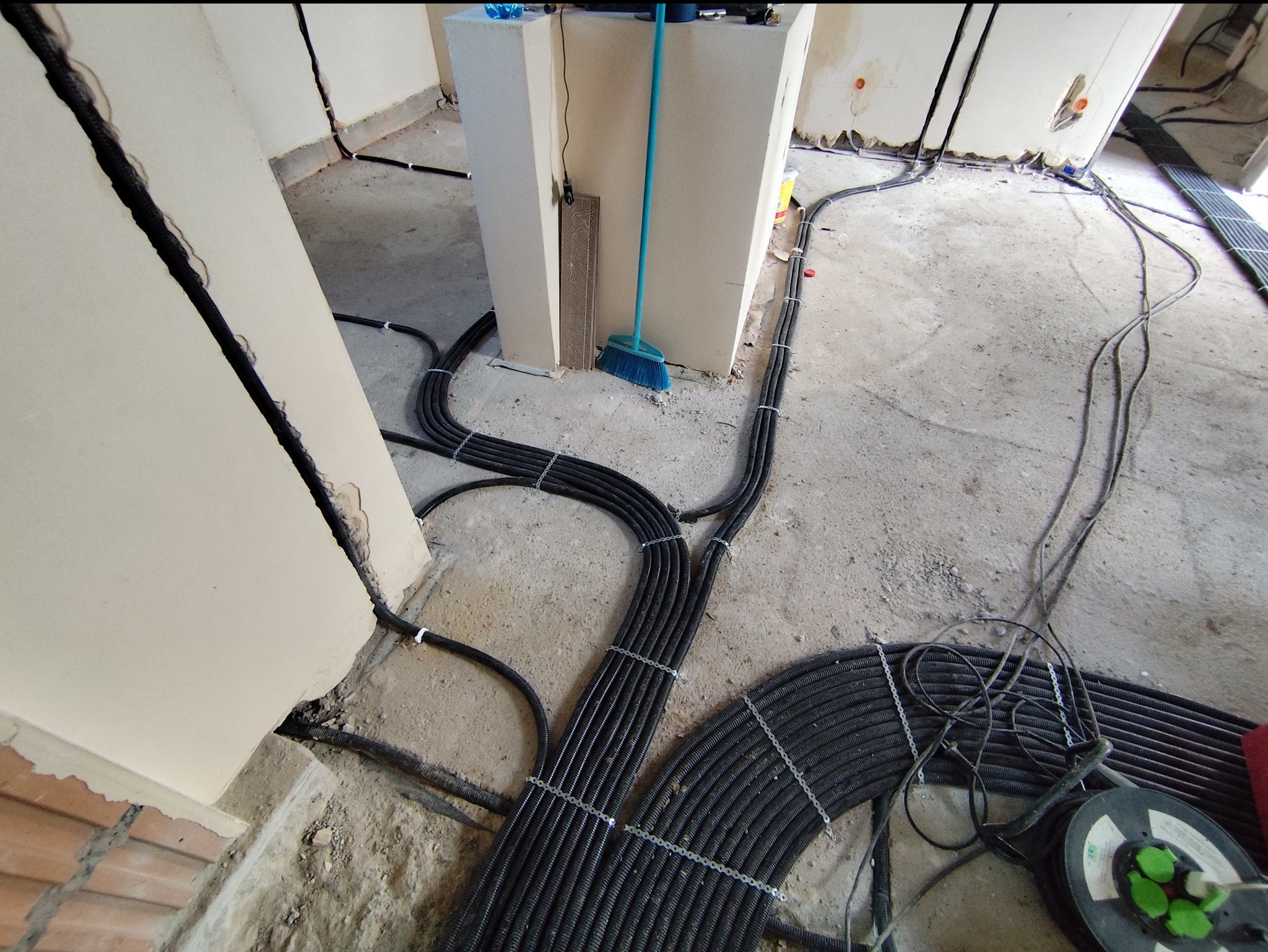 Rozprowadzenie czarnych rur karbowanych z przewodami elektrycznymi w surowym stanie deweloperskim, mocowanych metalowymi spinkami, widoczna miotła i zwój kabla na szpuli.
