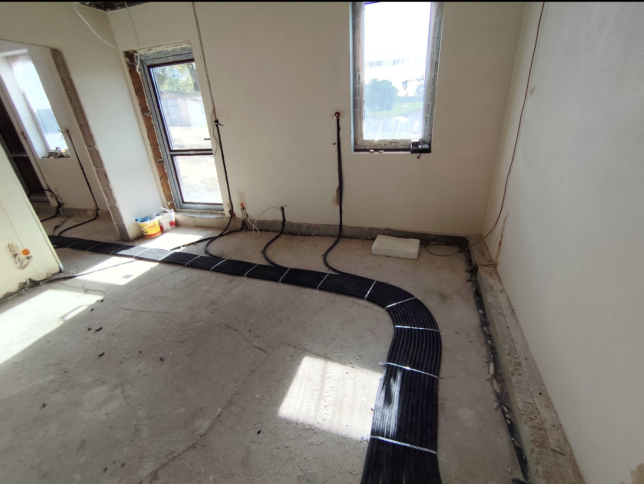 Rzędy czarnych rur w peszlu, tymczasowo spięte łańcuszkami, ułożone na surowym betonie podłogi w trakcie prac instalacyjnych w nowym budynku, widoczne okna i wystające przewody elektryczne ze ścian.