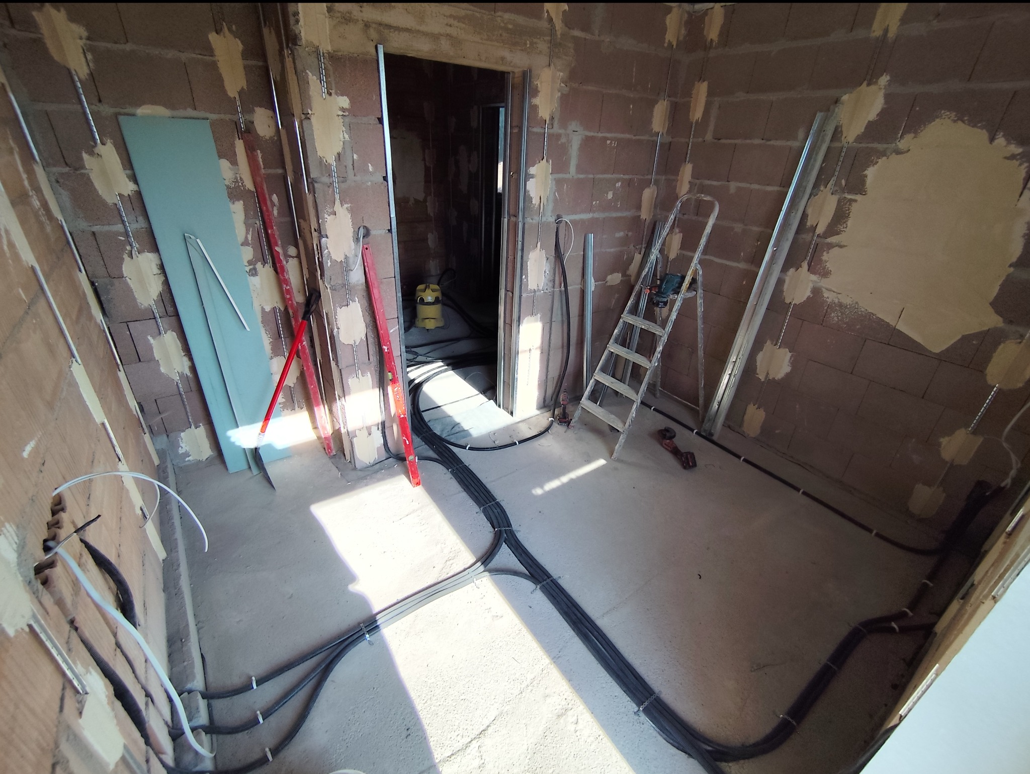 Wnętrze w trakcie remontu z widocznymi przewodami elektrycznymi ułożonymi na podłodze, poziomice oparta o ścianę, drabina rozstawiona w rogu pomieszczenia, płyta gipsowa oparta o ścianę...