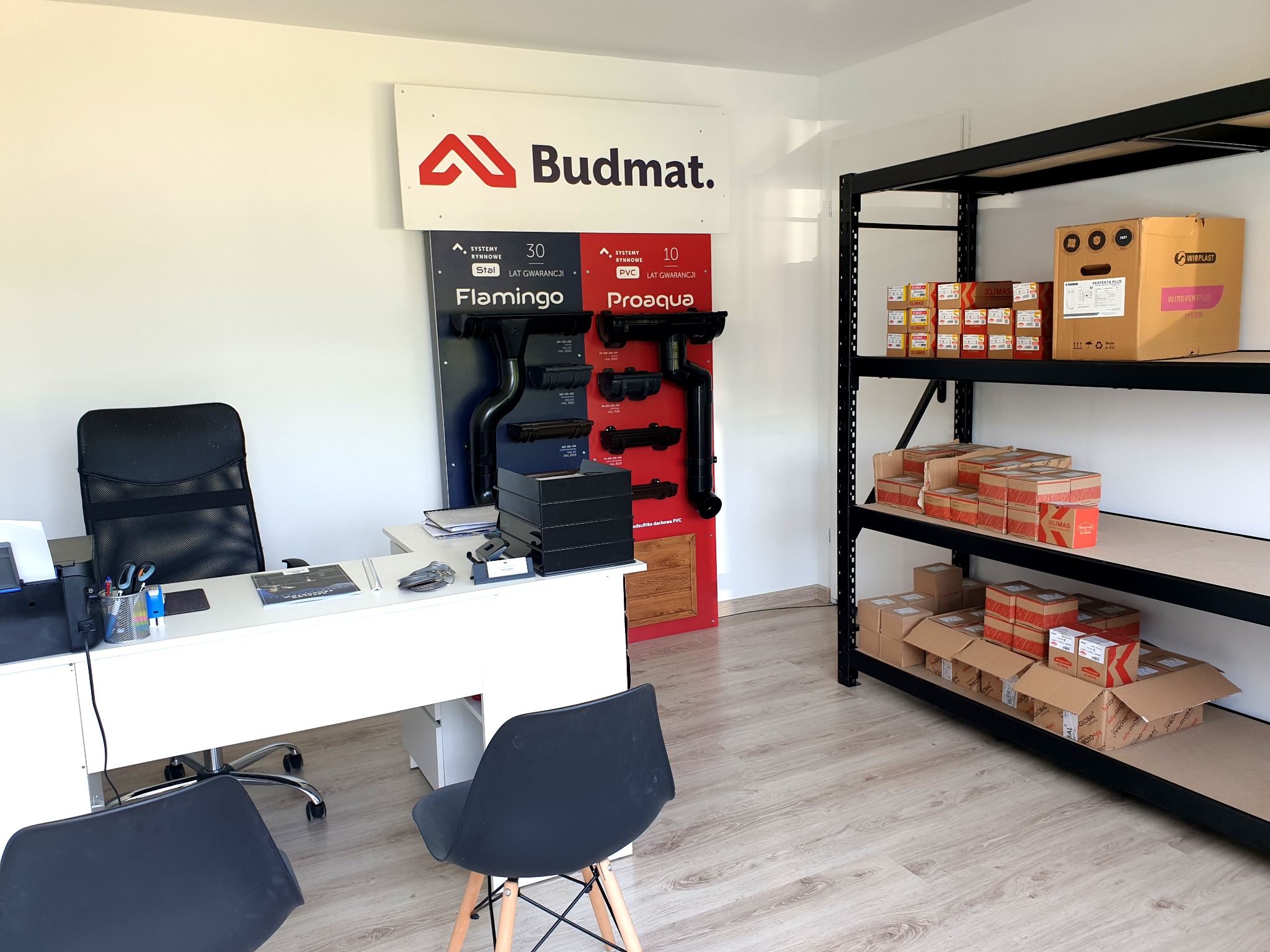 Biuro przedstawiciela firmy Budmat z ekspozycją systemów rynnowych Flamingo i Proaqua na ścianie oraz regałem z zapasem akcesoriów montażowych w kartonach.