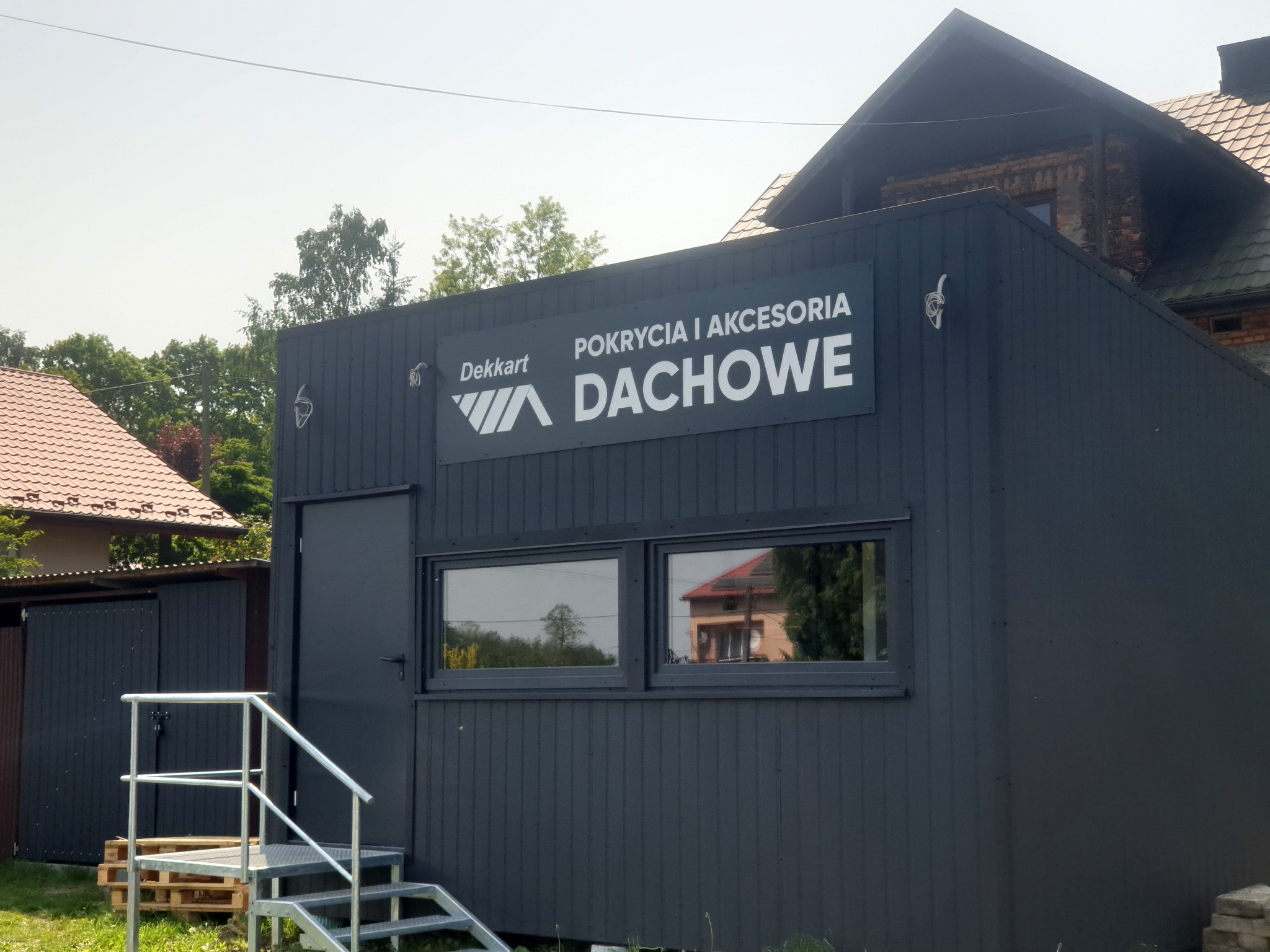 Budynek biura firmy Dekkart z napisem 'Pokrycia i akcesoria dachowe', widoczne schody wejściowe i okna z odbiciem sąsiedniego domu.