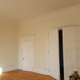 Jasne wnętrze po remoncie, ściany pomalowane na kremowo, widoczne drewniane podłogi, dwoje białych drzwi z ozdobnymi futrynami, listwa dekoracyjna pod sufitem i maszyna do cyklinowania parkietu.
