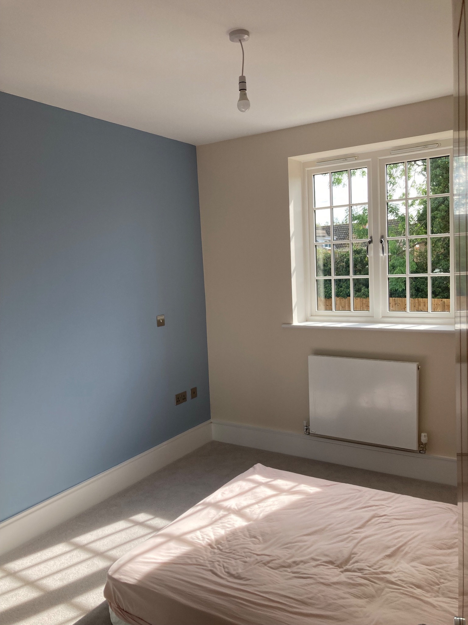 Pokój po malowaniu z jedną ścianą w odcieniach błękitu, jasną podłogą i grzejnikiem pod oknem, widoczne gniazdka elektryczne.