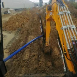 Żółta koparka JCB w trakcie wykonywania wykopu pod rurę wodociągową; widoczny fragment rury z niebieskiego tworzywa w wykopie oraz budynek magazynowy w tle.