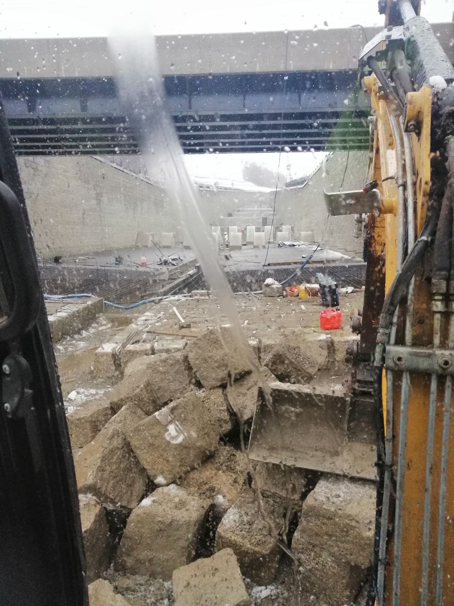 Widok z kabiny koparki na plac budowy z betonowymi blokami, zbrojeniami i konstrukcją mostu w tle podczas opadów śniegu.