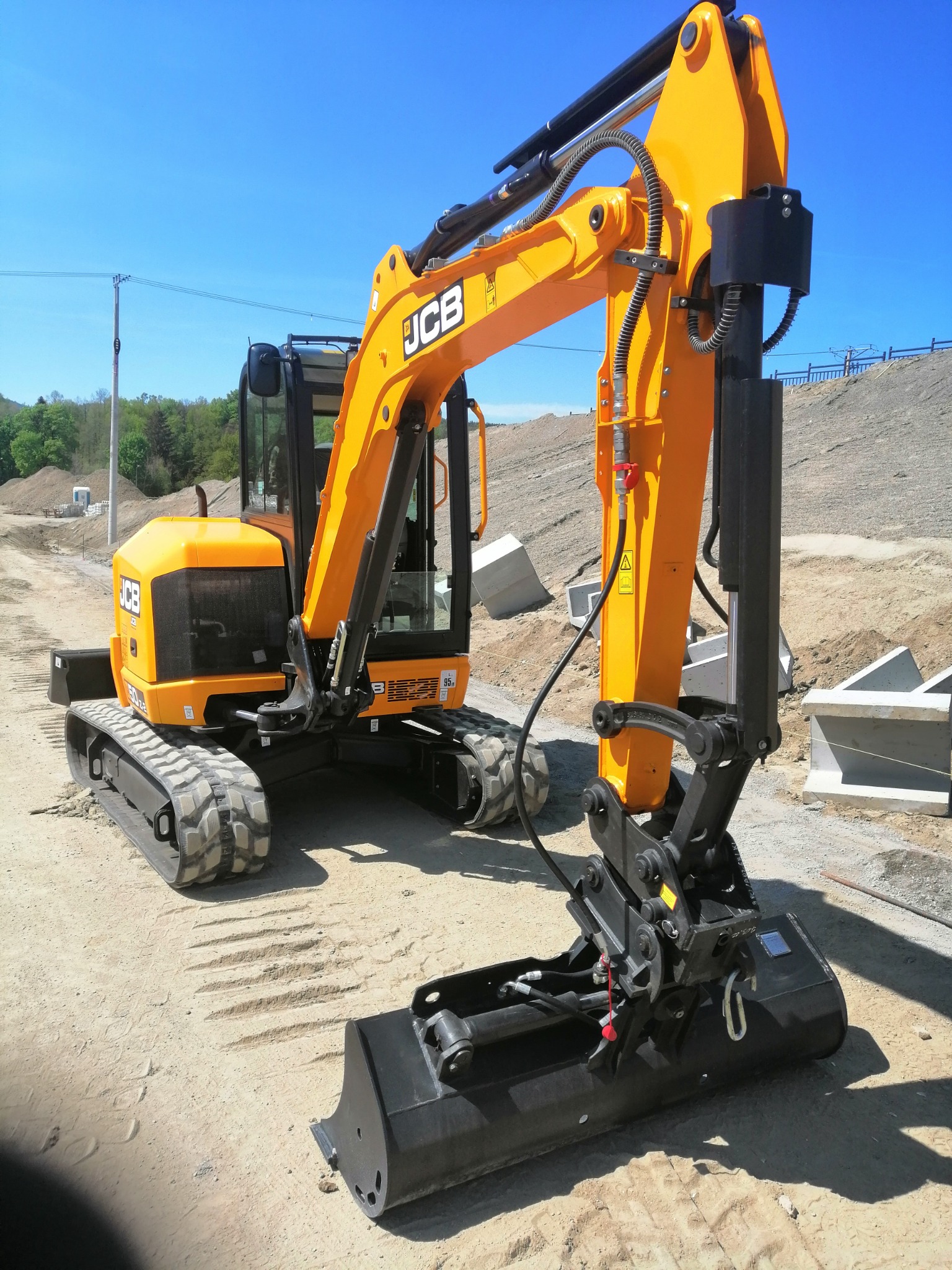 Żółta koparka gąsienicowa marki JCB z czarnym osprzętem hydraulicznym, stojąca na piaszczystym terenie budowy z widocznymi betonowymi prefabrykatami w tle, pod błękitnym niebem.