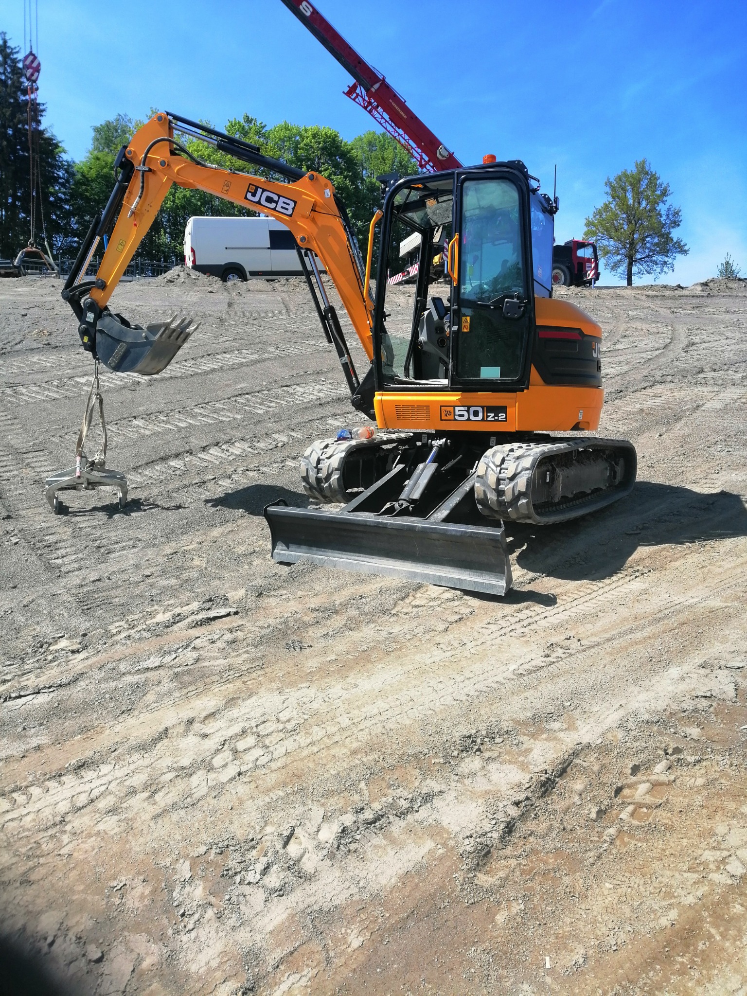 Żółto-czarna mini koparka JCB 50Z-2 na nierównym, szarym terenie z widocznymi śladami gąsienic, z podwieszonym elementem do podnoszenia ciężarów, dźwigiem w tle i drzewami na horyzoncie...
