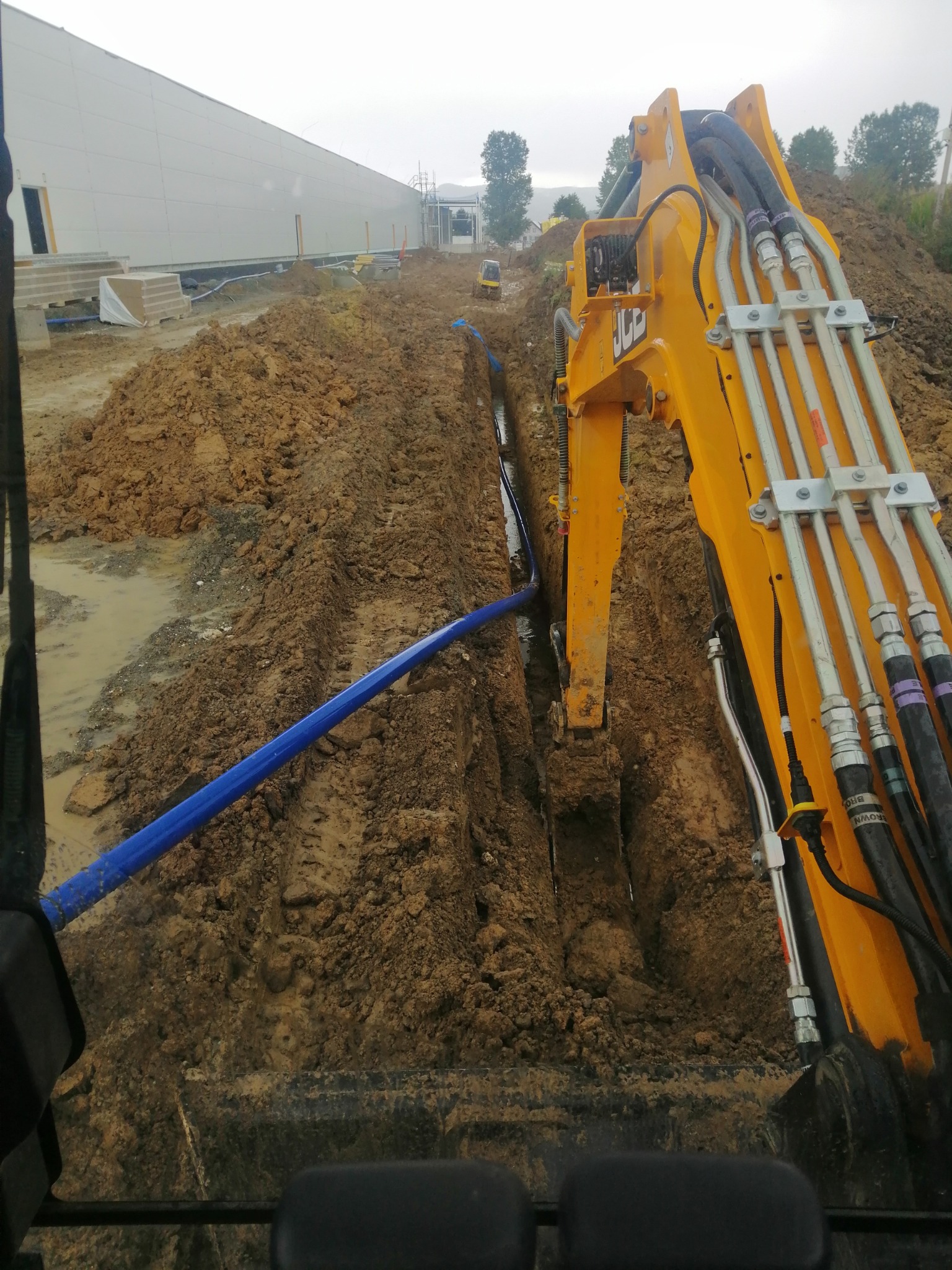 Żółta koparka JCB w trakcie wykonywania wykopu pod rurę wodociągową; widoczny fragment rury z niebieskiego tworzywa w wykopie oraz budynek magazynowy w tle.